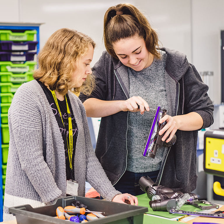 Two female students taking apart a Dyson vacuum cleaner head with a screwdriver.